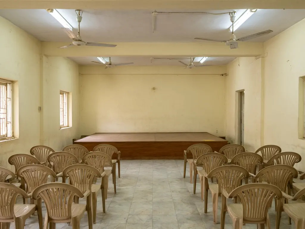 Marriage hall interior design result: dated cream hall with plastic chairs refitted in maroon and gold with carved arches, a golden false ceiling with crystal chandeliers, drapes, gold covered tables and a stage with a red sofa before AI redesign