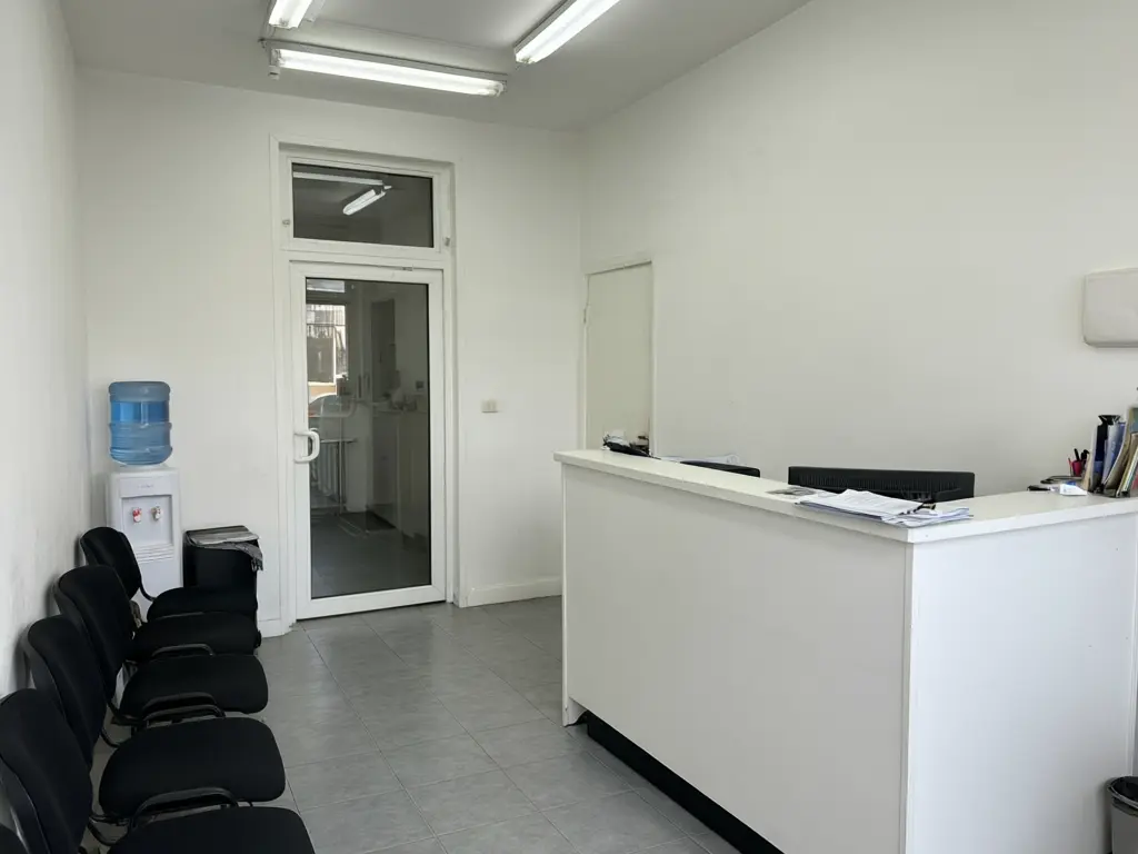Dental clinic reception design result: plain white reception with black chairs refitted with a curved white and oak desk, a lit mint green tooth logo panel, soft green waiting chairs, an oak slat wall and a terrazzo floor before AI redesign